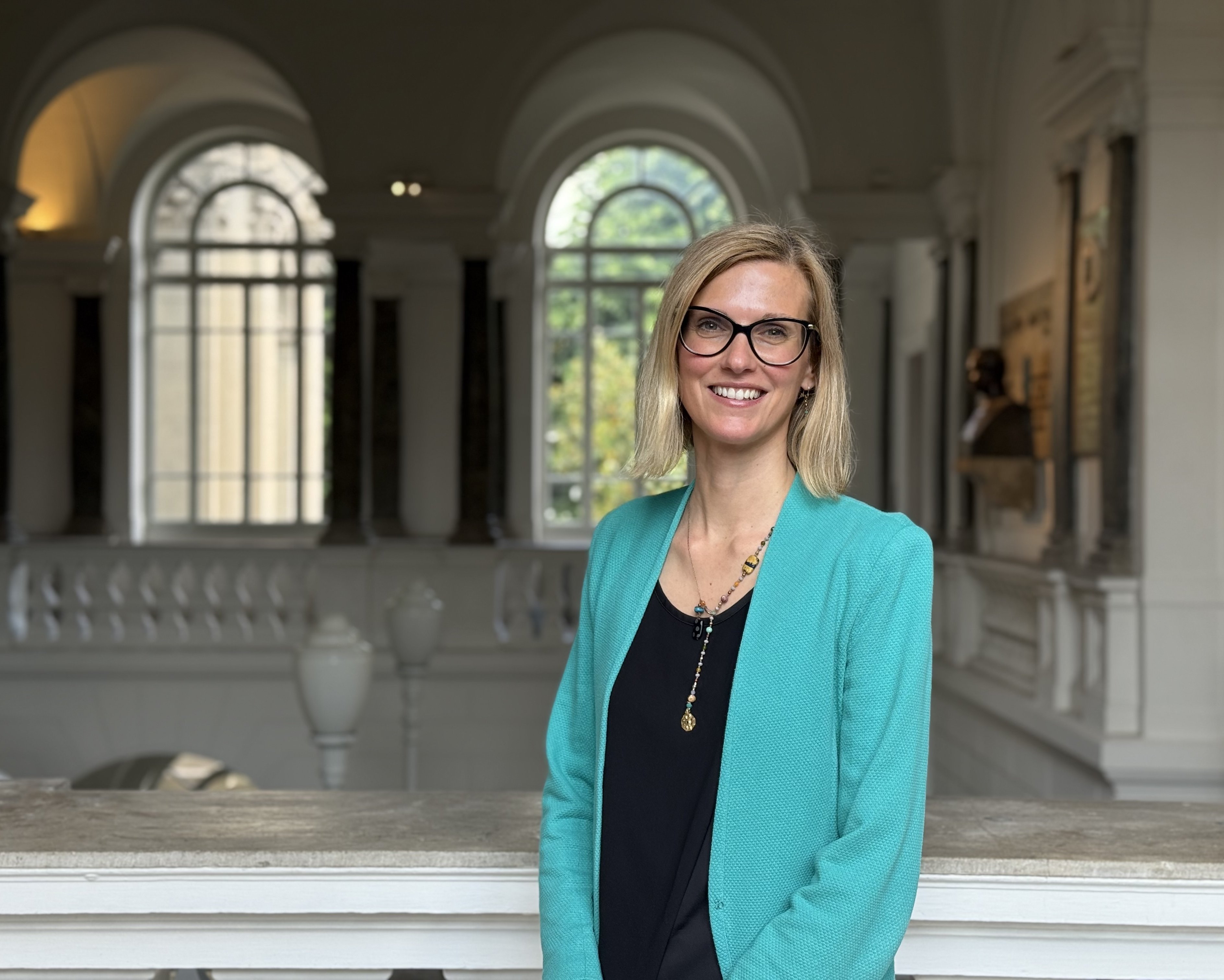 Donna bionda con capelli corti e occhi azzurri. Indossa un paio di occhiali con montatura nera, una giacca color tiffany e una camicia nera. Sullo sfondo, finestre ad arco e balconata interna al Rettorato. 